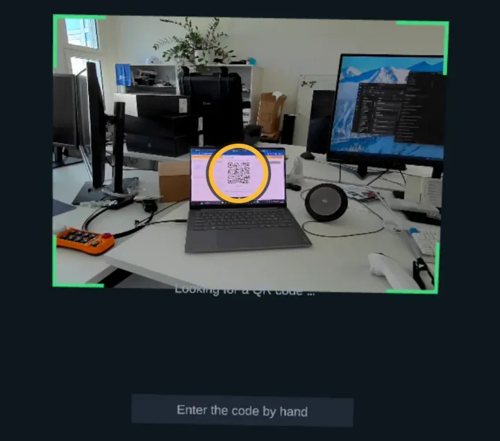 Das VR-Headset zeigt das Bild seiner Kamera in einem grünen Rahmen: ein Schreibtisch mit einem Laptop, auf dem der QR-Code aus dem Dashboard steht. Darunter der Knopf «Enter the code by hand». Markiert: der QR-Code auf dem Laptop.